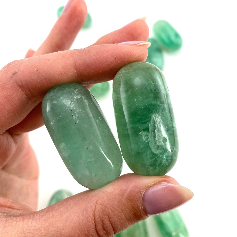 May include: Two smooth, green fluorite stones, polished to a high shine, are held in a hand. The stones are oval shaped and have a translucent appearance.