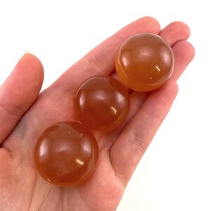 May include: Three smooth, round, orange carnelian spheres in a hand. The spheres are polished and have a translucent quality.