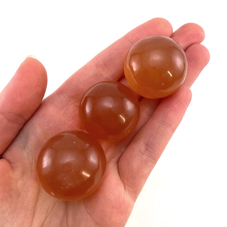 May include: Three smooth, round, orange carnelian spheres in a hand. The spheres are polished and have a translucent quality.