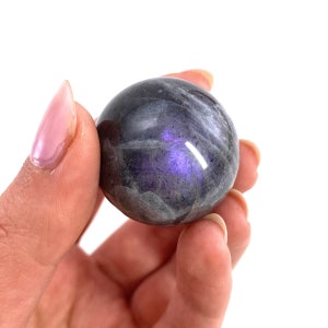 May include: A polished, spherical gemstone with a dark gray base color and iridescent purple and blue flashes. The stone is held in a hand against a white background, showcasing its smooth, reflective surface. The gemstone is a decorative item.