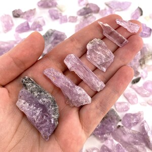 K&ouml;nnte beinhalten: Eine Sammlung von rosa und violetten Lepidolith-Kristallspitzen und rohen St&uuml;cken. Die Kristalle sind in verschiedenen Gr&ouml;&szlig;en und Formen, einige haben ein undurchsichtigeres Aussehen als andere.