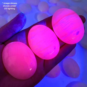 May include: Three oval stones glowing bright pink under UV light, held in a hand. The stones have a smooth surface with subtle patterns. The background shows other stones and a blue glow.