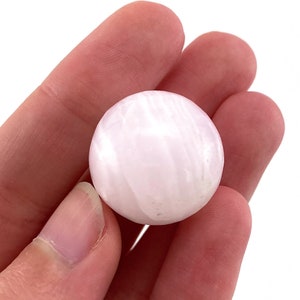 May include: A smooth, white, and pink sphere of selenite crystal. The sphere is polished and has a striated pattern.