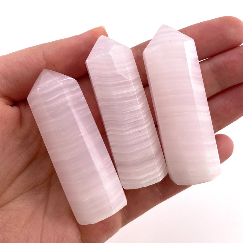 May include: Three pink selenite wands with a smooth, polished finish and striated patterns. The wands are arranged in a row, with the tips pointing towards the right.