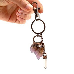 May include: A copper-toned keychain with a large ring and a lobster clasp. The keychain features a cluster of purple amethyst crystals and a small, clear quartz crystal pendant. The crystals are attached with a chain.