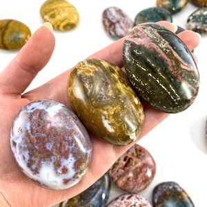 May include: A hand holding several polished oval stones. The stones display a variety of colours, including white, brown, green, and maroon. The stones have a smooth, glossy finish and are likely used for decorative or therapeutic purposes.