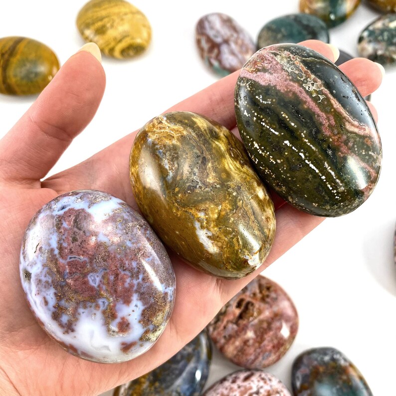 May include: A hand holding several polished oval stones. The stones display a variety of colours, including white, brown, green, and maroon. The stones have a smooth, glossy finish and are likely used for decorative or therapeutic purposes.