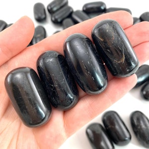 Op de afbeelding: Vier gladde, zwarte, getrommelde stenen, waarschijnlijk obsidiaan of zwarte toermalijn, worden in een hand gehouden. De stenen zijn ovaalvormig en hebben een glanzende afwerking.