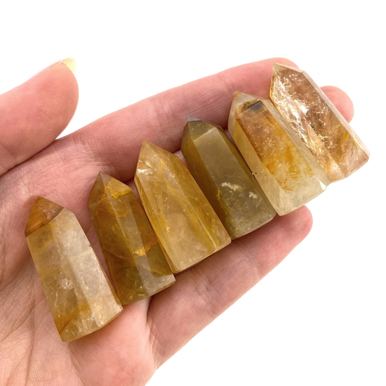 May include: Six golden quartz crystal points, each with a unique pattern and color variation. The crystals are held in a hand, showcasing their natural beauty and varying shades of yellow and brown.