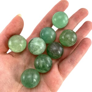 May include: A collection of eight green fluorite spheres, each with a smooth, polished surface. The spheres are arranged in a hand, showcasing their size and color.