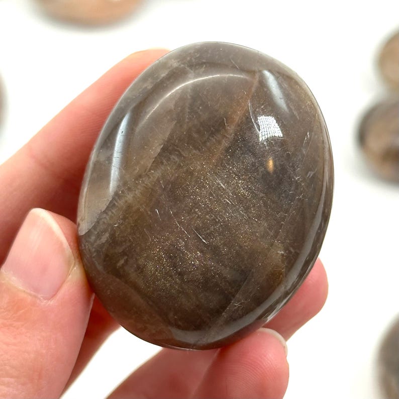 May include: A polished, oval-shaped stone with a shimmering, golden-brown appearance. The stone has a smooth, reflective surface with subtle streaks and variations in color, creating a natural, earthy aesthetic. The stone is held in a hand.