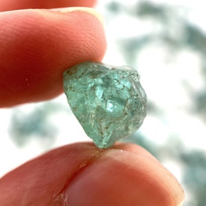 May include: A single, rough-cut aquamarine gemstone with a light blue-green color. The gemstone is translucent and has a slightly cloudy appearance.