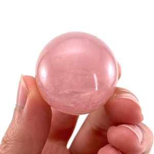 May include: A smooth, polished rose quartz sphere, a light pink color with subtle white veining. The sphere is being held in a hand.