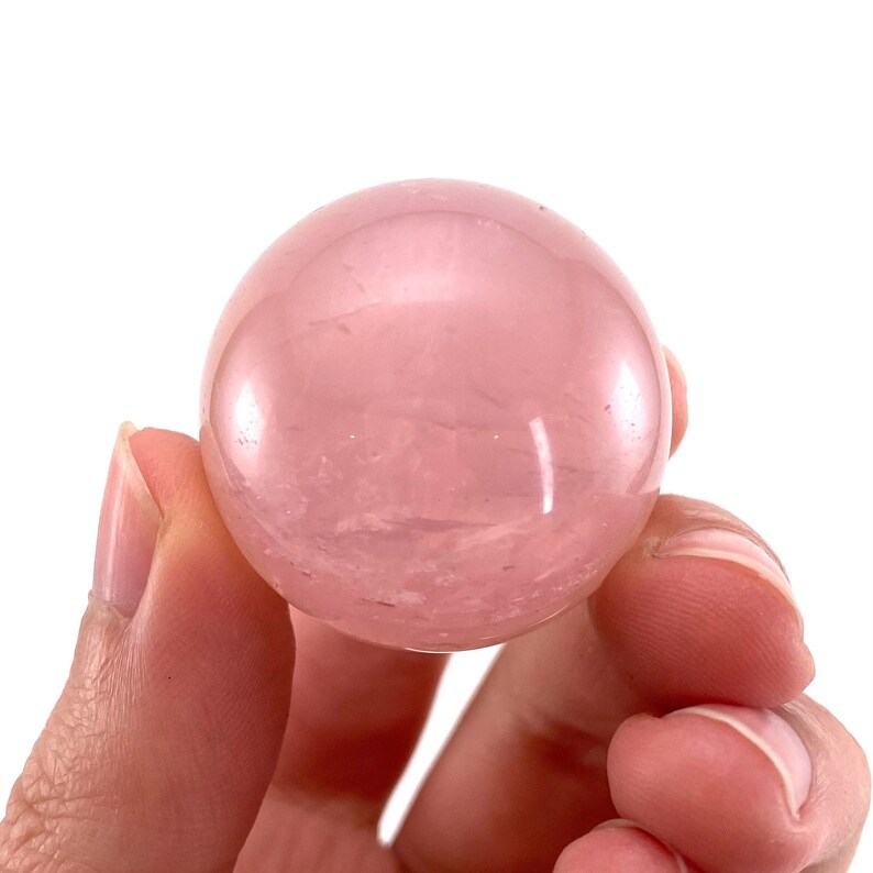 May include: A smooth, polished rose quartz sphere, a light pink color with subtle white veining. The sphere is being held in a hand.