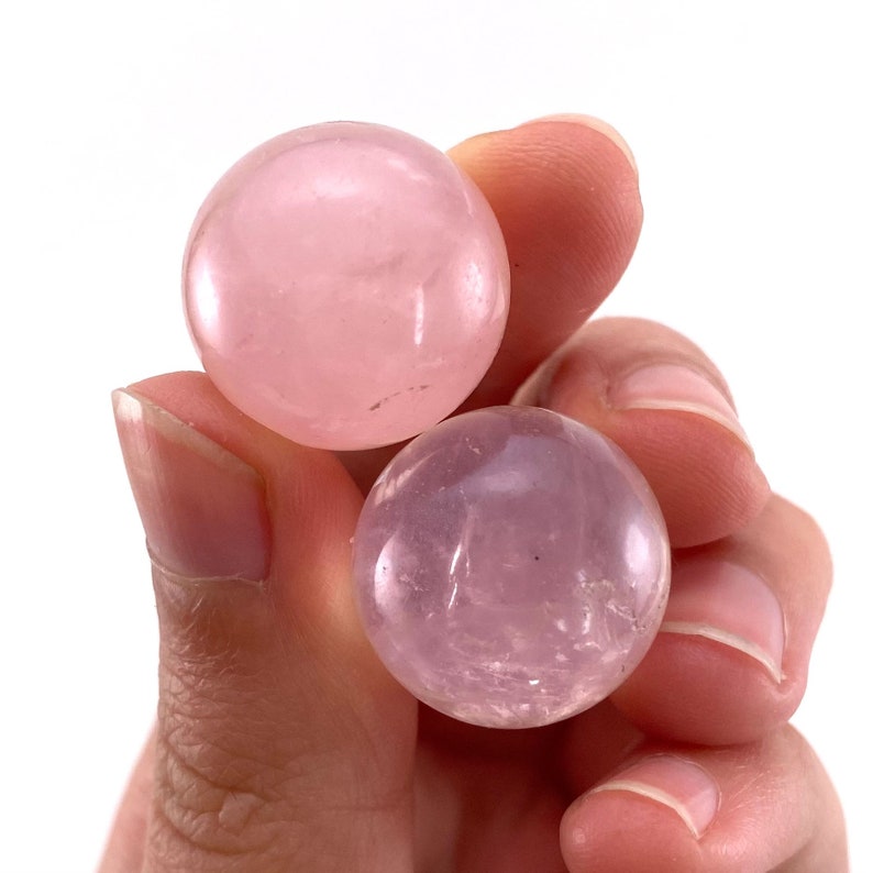 May include: Two rose quartz spheres, one a lighter pink than the other, are held in a hand against a white background. The spheres are smooth and translucent, showcasing the natural colour variations of the stone.