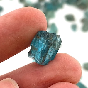May include: A teal blue gemstone with a rough, faceted surface. The gemstone is held in a hand.