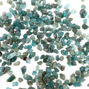 May include: A collection of teal blue apatite crystals, scattered on a white background. The crystals are various sizes and shapes, with some having a more pointed shape and others being more rounded.