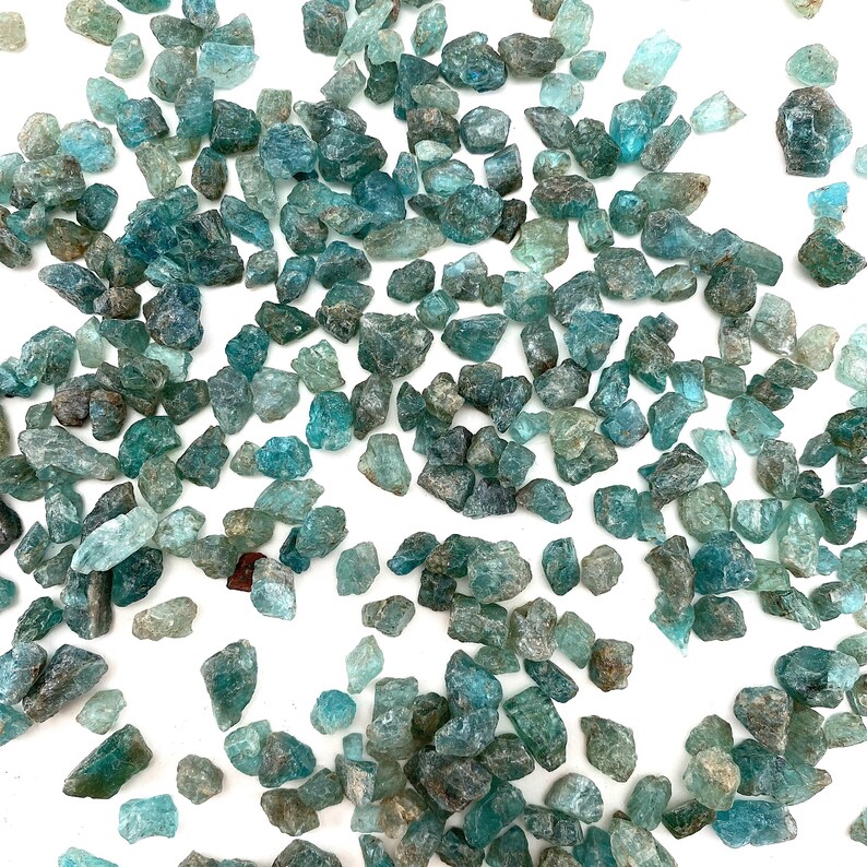 May include: A collection of teal blue apatite crystals, scattered on a white background. The crystals are various sizes and shapes, with some having a more pointed shape and others being more rounded.