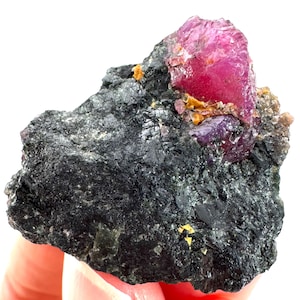 以下が含まれることがあります： 生のルビー結晶クラスター。深い赤色のルビーは、金やその他の鉱物の斑点がある濃い灰色の岩のマトリックスにセットされています。石の粗い質感が目に見えます。