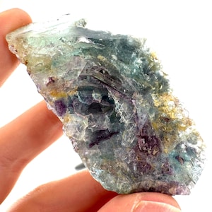May include: A translucent, multi-colored fluorite specimen. The stone displays a mix of green, purple, and yellow hues, with a crystalline structure. The fluorite is held in a hand against a white background.