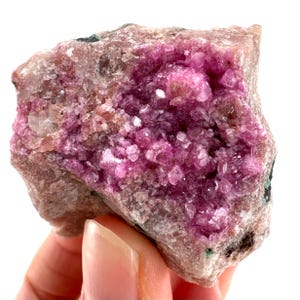 May include: A natural mineral specimen with a cluster of bright pink crystals. The crystals are set within a matrix of light brown and tan rock. The overall appearance is rough and textured, with a mix of colours and shapes.