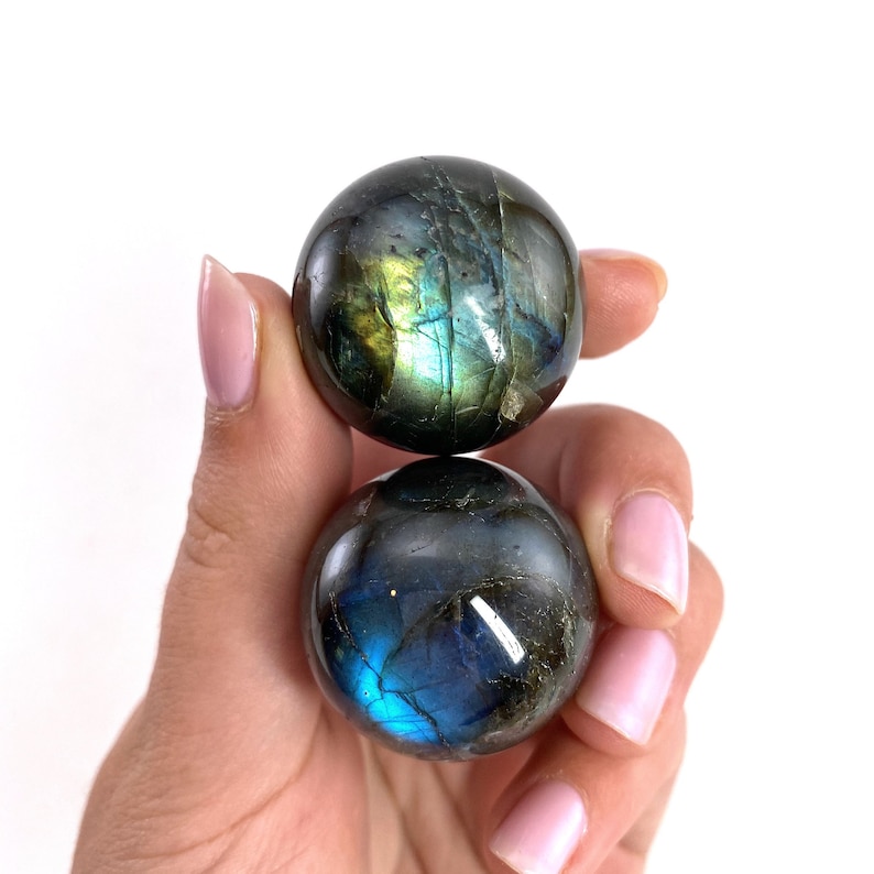 May include: Two polished labradorite spheres, held in a hand, display iridescent flashes of blue, green, and gold. The spheres have a smooth, rounded shape and a dark, speckled appearance, reflecting light.