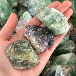 K&ouml;nnte beinhalten: Eine Sammlung von gr&uuml;nen, violetten und schwarzen Fluoritkristallen. Die Kristalle sind roh und haben ein nat&uuml;rliches, unpoliertes Aussehen.