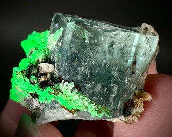 May include: A large, clear, green fluorite crystal with a cubic shape. The crystal is partially covered in a green mineral, possibly epidote. The crystal is sitting on a bed of white and gray matrix.