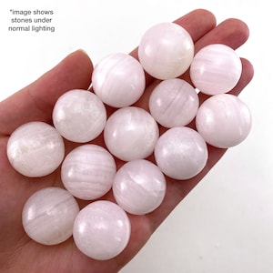 May include: A collection of 12 smooth, white, and pink selenite spheres. The spheres are arranged in a hand, with some overlapping.