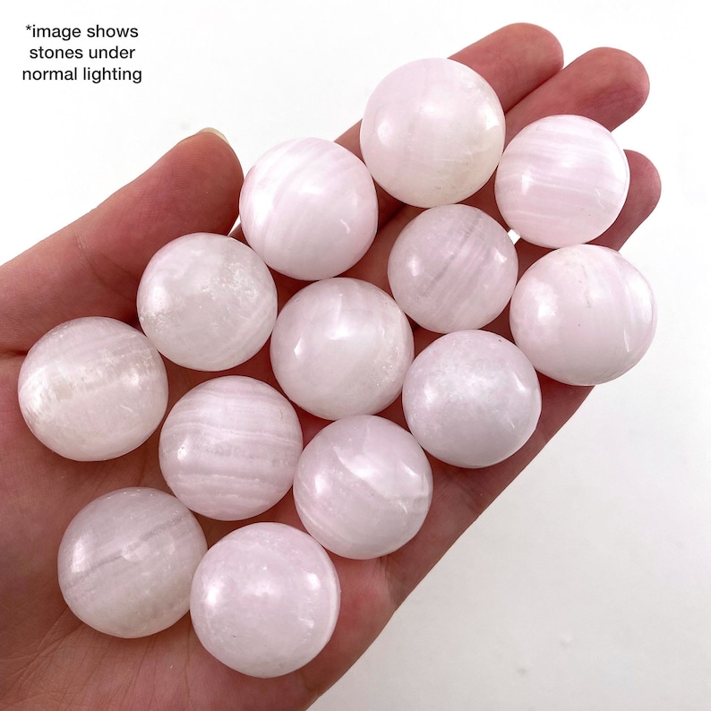 May include: A collection of 12 smooth, white, and pink selenite spheres. The spheres are arranged in a hand, with some overlapping.