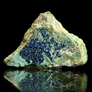 May include: A close-up of a mineral specimen featuring a rough, textured surface. The stone is primarily green with clusters of deep blue crystals. The dark background enhances the colors and textures of the rock.