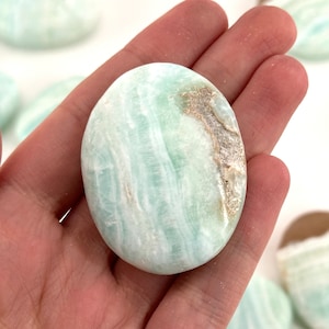 Op de afbeelding: Een gladde, lichtgroene en witte gepolijste celestijn palmstone. De steen heeft een draaiende structuur en een licht ruwe textuur.