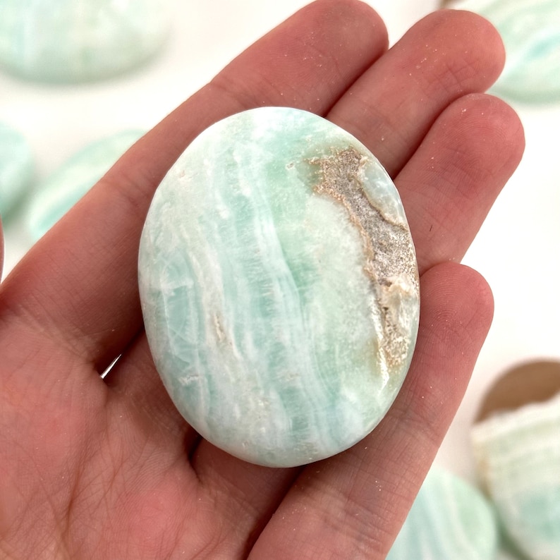 Op de afbeelding: Een gladde, lichtgroene en witte gepolijste celestijn palmstone. De steen heeft een draaiende structuur en een licht ruwe textuur.