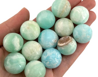 May include: A hand holds a collection of twelve spherical stones. The stones vary in shades of blue-green, with some displaying white veining and a few having brown patches. The spheres are smooth and appear to be polished gemstones.