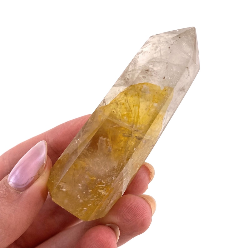 May include: A clear quartz crystal point with golden inclusions. The crystal is held in a hand and has a polished, faceted shape. The golden inclusions add a unique visual texture to the crystal.