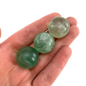 May include: Three green fluorite spheres, polished smooth, with a translucent, almost glassy appearance. The spheres are arranged in a row on a hand.