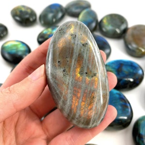 K&ouml;nnte beinhalten: Ein glatter, ovaler Labradorit-Handstein mit schillernden blauen, gr&uuml;nen und goldenen Farben. Der Stein wird in einer Hand gehalten.