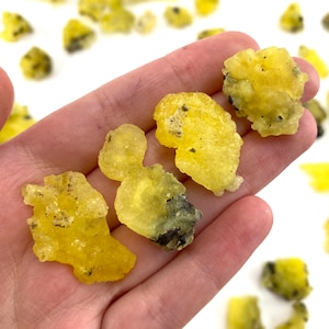 May include: Four yellow and black mineral specimens, also known as yellow calcite, on a hand. The mineral specimens are irregularly shaped and have a rough, crystalline texture.