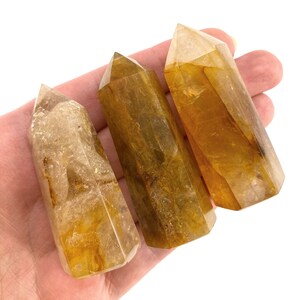 May include: Three golden-yellow quartz crystal points with a polished, faceted design. The crystals vary in size and have a translucent quality, with visible internal patterns. The crystals are held in a hand against a white background.