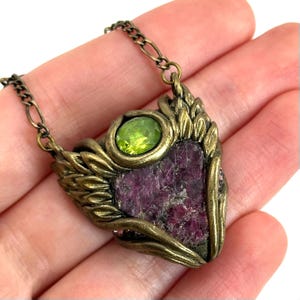 May include: A gold pendant necklace with a large, rough, purple gemstone set in a gold frame. The pendant is surrounded by gold wings and a smaller, round, green gemstone.