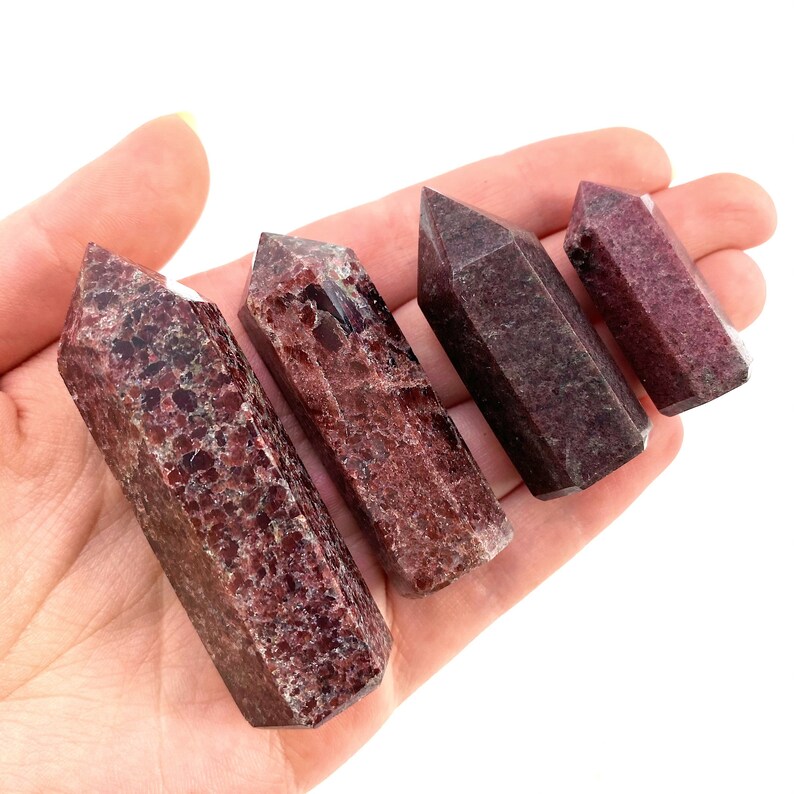 May include: Four red and brown gemstone points, also known as red and brown crystal wands, are arranged in a row on a hand. The points are different sizes and have a natural, rough texture.