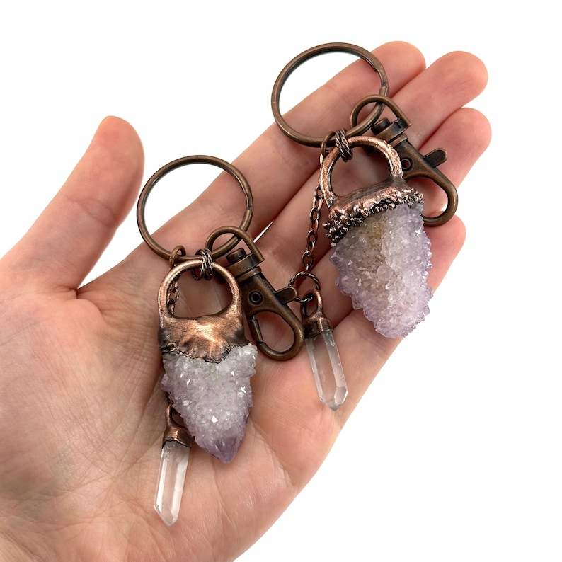 May include: Two keychains with amethyst crystal clusters and clear quartz pendants. The keychains have copper-colored metal accents and clasps. The crystals are a pale purple color.
