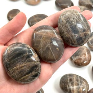 May include: Several polished, oval-shaped stones in shades of brown and tan are held in a hand. The stones have a smooth, glossy surface with natural patterns and variations in color. The stones are a natural product.