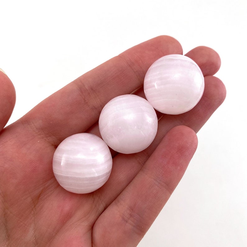 May include: Three smooth, white selenite spheres with a subtle pink hue. The spheres are polished and have a striated pattern.