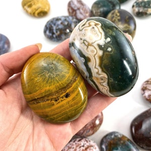 May include: Two polished oval-shaped stones. One is a green, white, and brown jasper with a swirling pattern. The other is a yellow and brown agate with a concentric pattern.
