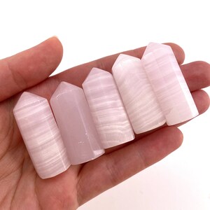 May include: Five white selenite crystal wands with a pink hue. The wands are smooth and polished, with a pointed tip and a flat base.
