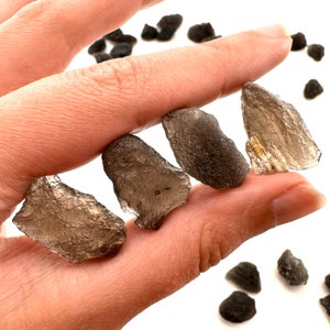 May include: Four smoky quartz crystals, each with a unique shape and size, are held in a hand. The crystals are translucent and have a dark grey colour.