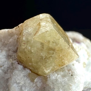 Może przedstawiać: Fasetowany, jasnożółty kryształ o geometrycznym kształcie, spoczywający na białej, teksturowanej podstawie mineralnej. Powierzchnia kryształu ma subtelne wariacje koloru i tekstury, na ciemnym tle.