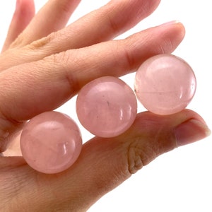 May include: Three polished rose quartz spheres, each with a soft pink hue. The spheres are smooth and round, showcasing the natural colour variations of the stone. The spheres are held in a hand.