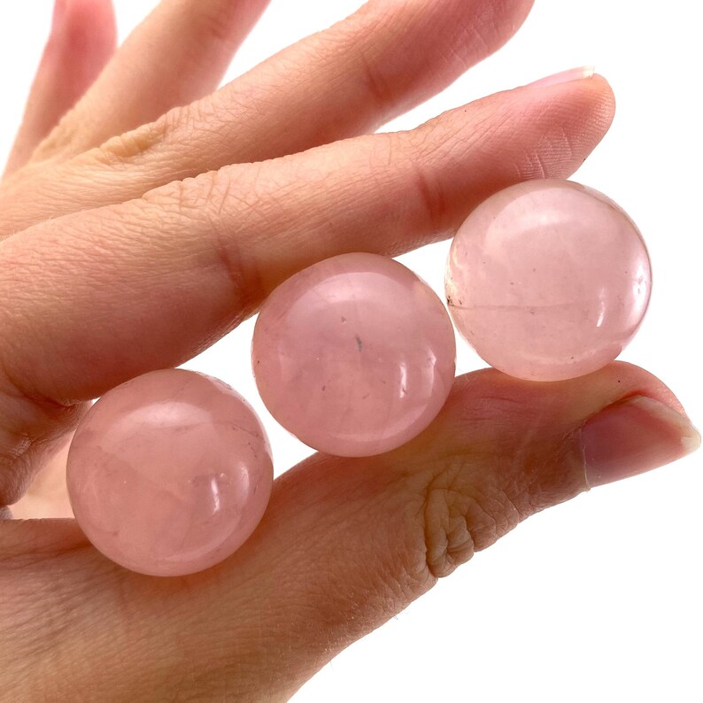 May include: Three polished rose quartz spheres, each with a soft pink hue. The spheres are smooth and round, showcasing the natural colour variations of the stone. The spheres are held in a hand.